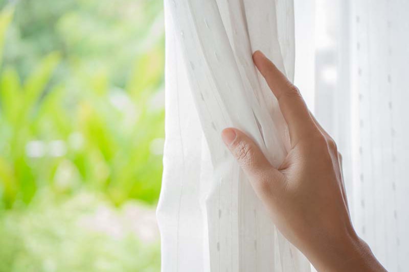 Woman's hand opening curtains in the bedroom with natural light and garden background., spring money-saving tips