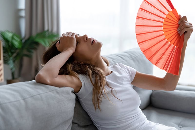 Why Is My AC Blowing Hot Air? Side view exhausted young biracial girl using paper waver, suffering from hot summer weather or high temperature at home. Unhappy overheated millennial mixed race woman feeling uncomfortable indoors.