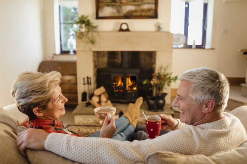 How Do I Keep My Heat During Extreme Cold Weather? - Mature couple sat infront of a fire drinking hot chocalate from mugs.