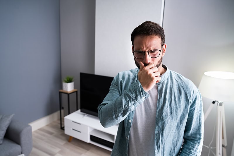 A man wearing glasses and a light blue button-down shirt over a gray t-shirt is standing in a living room.