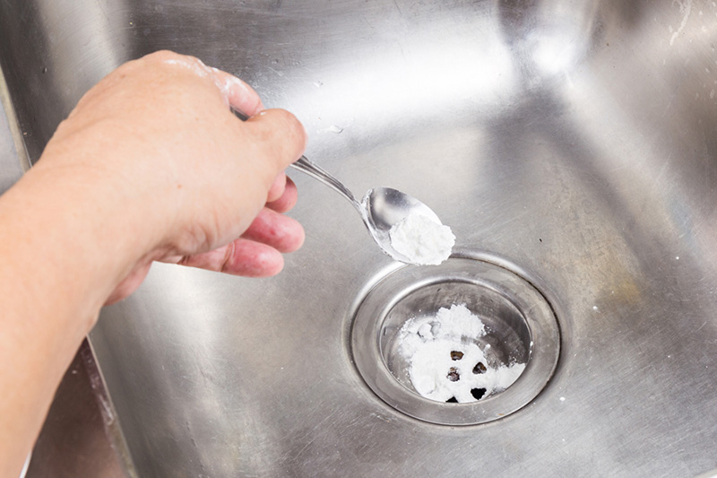 How to Unclog a Drain. A photo of a person's hand holding a spoon, sprinkling baking soda into a clogged sink drain.